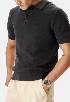 Homme portant un polo en maille à manches courtes de couleur foncée, pantalon de couleur claire, et une montre-bracelet argentée avec un cadran noir, mains dans les poches.