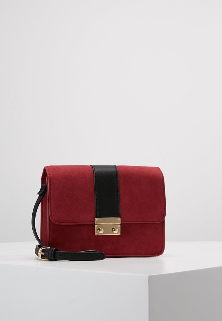 Sac à main en daim rouge avec une bandoulière en cuir noir, doté d'une boucle dorée et d'une forme rectangulaire, à la texture lisse et au design minimaliste.