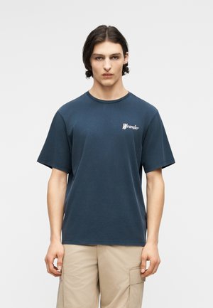 Νεαρός άνδρας με μεσαίου μήκους σκούρα μαλλιά φοράει μπλε ναυτικό μπλουζάκι Wrangler και μπεζ παντελόνι cargo, κοιτάζοντας ευθεία μπροστά με φόντο απλή επιφάνεια.