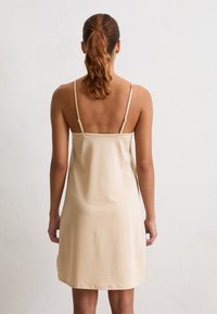 Donna con capelli castani mossi legati in una coda di cavallo, che indossa un vestito satinato beige a sottoveste con spalline sottili e regolabili, in piedi contro un muro bianco semplice.