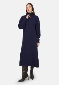 Abito maglione blu navy con un collo alto, maniche oversize e una vestibilità rilassata, abbinato a stivali marroni al ginocchio con punta affusolata.