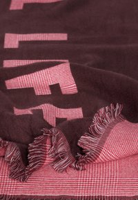 Mørk maroon og lyserød tæppe med et tekstureret mønster, der har frynsekanter og et skakmønster. Stoffet ser blødt og tykt ud.