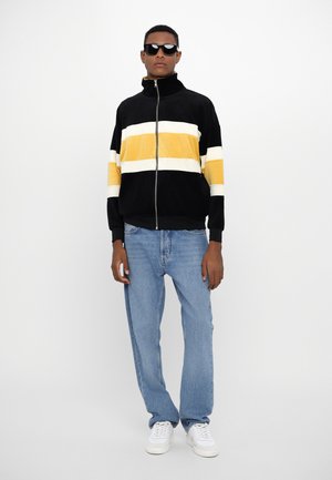 Black zippered jacket with yellow and white horizontal stripes, paired with light blue straight-leg jeans and white sneakers. Model wearing sunglasses.