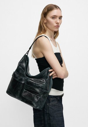 Junge Frau trägt ärmelloses Oberteil und Jeans und hält große schwarze Leder-Umhängetasche mit mehreren Fronttaschen.