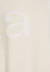 Kremowa tkanina o miękkiej fakturze, na której widoczna jest duża, subtelna biała nadrukowana litera. Na boku widoczny jest detal szwu.