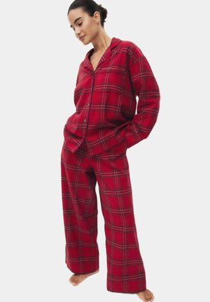 Mujer con pijama suelto de cuadros rojos con mangas largas y pantalones de pierna ancha, de pie descalza frente a un fondo liso.