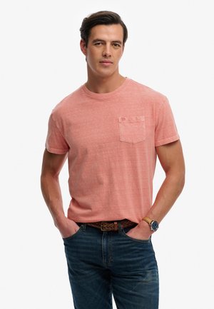 Mand iført laksesfarvet kortærmet T-shirt med brystlomme, blå jeans, brun flettet bælte og armbåndsur, stående med hænderne i lommerne.