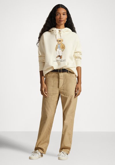 Mulher com cabelo comprido e encaracolado, vestindo um hoodie creme com gráfico de urso, calças bege, cinto preto e ténis brancos, em pé contra um fundo branco.