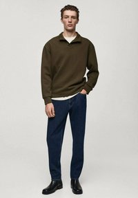 Pullover verde scuro con colletto alto, abbinato a jeans blu a gamba dritta e stivali neri all'altezza della caviglia, su uno sfondo neutro.