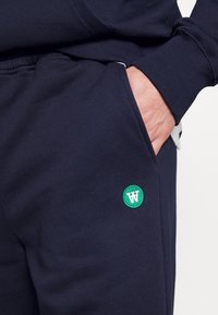 Pantalones de chándal navy hechos de una mezcla de algodón, cuentan con un bolsillo lateral y un parche de logo circular verde con texto blanco en la zona de la cadera.