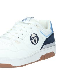 Vit sneaker med marinblåa och ljusblåa accenter, med en texturerad ovandel, perforerade detaljer och en gummisul. Logotypen är tryckt på sidan.