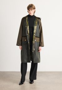 Olijfgroene lange jas met een capuchon, voorzien van een geruite voering en gouden hardware accenten. Draagt over een zwarte coltrui en zwarte broeken.