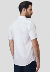 Biała koszula z krótkim rękawem w granatowe kropki, z kołnierzykiem zapinanym na guziki i zaokrąglonym dołem. Gładka faktura materiału i dopasowany fason.