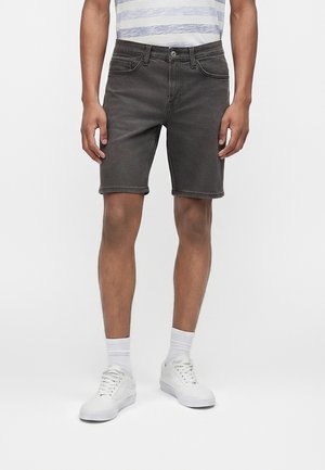 Osoba u tamnosivim traper kratkim hlačama, bijelim čarapama, bijelim tenisicama i bijelo-sivoj prugastoj majici, stoji na jednobojnoj pozadini.