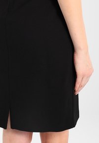 Mustassa mekossa on sivuaukko ja se on valmistettu sileästä kankaasta. Lähikuva, jossa näkyy osa mekosta ja käsi, joka lepää sen päällä.