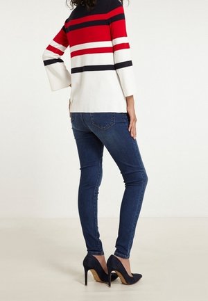 Kobieta ubrana w ciemnoniebieskie obcisłe dżinsy, czarne buty na wysokim obcasie i sweter z długim rękawem w poziome paski w kolorach czerwonym, czarnym i białym, zwrócona tyłem.