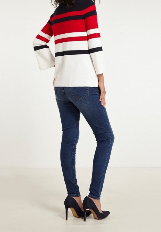 Femme portant un jean skinny bleu foncé, des chaussures à talons noirs et un pull à manches longues avec des rayures horizontales rouges, noires et blanches, vue de dos.