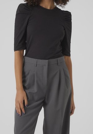 Femme portant un haut noir à manches bouffantes et un pantalon gris taille haute plissé, debout avec les bras relâchés le long du corps.