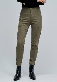 Pantalon cargo vert olive à coupe ajustée, avec des poches latérales, une fermeture à bouton et une texture de tissu lisse. Agencé avec des bottines noires.