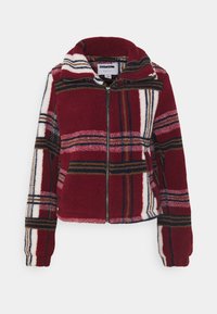 Veste polaire à carreaux rouges et blancs ; elle présente un col montant, une fermeture éclair complète et des manches longues avec des accents noirs et marron dans le motif.