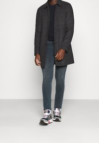 Sort ternet frakke med krave over en navy skjorte, kombineret med mørke jeans og hvide New Balance-sneakers med røde og blå detaljer.