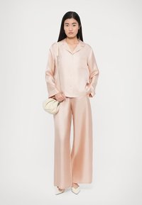 Satin-Pyjama-Set in zartem Rosa, bestehend aus einem Hemd mit Knöpfen und Tasche sowie weitem Hosenbein. Dazu passende cremefarbene Tasche und High Heels.