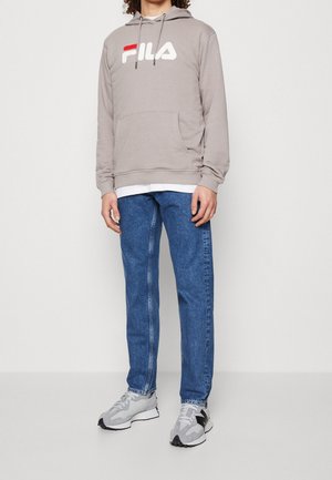 Persona con sudadera con capucha beige de FILA, jeans azules, camiseta blanca y zapatillas gris-negras, de pie contra una pared lisa y clara.