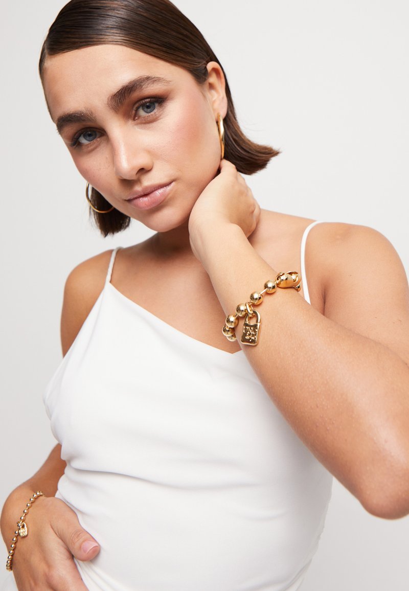 Joven con cabello corto y castaño, vistiendo una blusa blanca sin mangas y pendientes de aro dorados, posando con pulseras de dijes dorados en ambas muñecas.