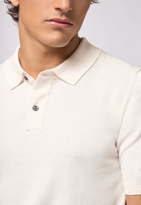Polo shirt color crema chiaro realizzato in morbido tessuto a maglia. Presenta un colletto classico, maniche corte e un dettaglio a un bottone sullo scollo.