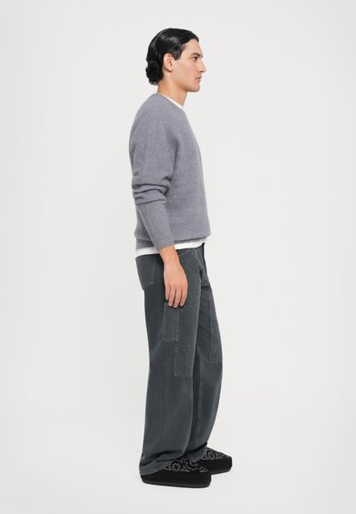 Grijze wollen trui, losse pasvorm, gedragen over een wit shirt; donkere cargobroek met zakdetails, en zwarte slippers met geborduurd ontwerp.