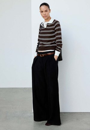 Jeune personne debout, les mains dans les poches, portant une chemise à manches longues rayée marron et un large pantalon noir devant un mur blanc.