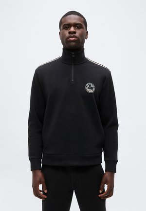 Uomo che indossa un maglione nero con una cerniera a quattro parti e un logo del club di tennis sul petto sinistro, in piedi contro uno sfondo semplice.