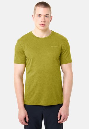 Giovane uomo che indossa una maglietta aderente a maniche corte verde oliva e pantaloni scuri, in piedi contro uno sfondo chiaro e semplice.