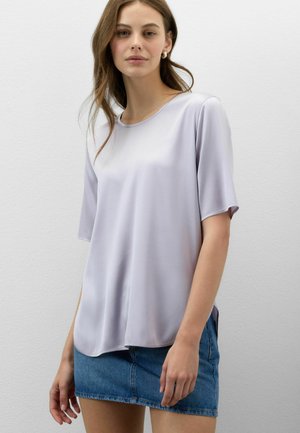Frau mit hellbraunen Haaren, die eine lockere, helllavendelfarbene kurzärmelige Bluse und einen blauen Jeans-Minirock vor einer einfarbigen Wand trägt.