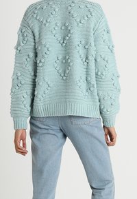 Ljusblå stickad tröja med strukturerade boblemönster, bärs av en person som vänder ryggen mot kameran, tillsammans med ljusa denimjeans.