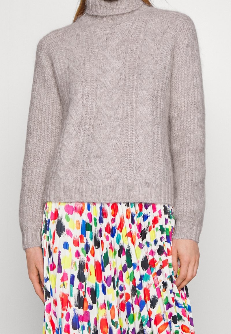 Lys grå strikket sweater med høj krave, prydet med et kabelstrikket design. Matchet med en flerfarvet mønstret nederdel.