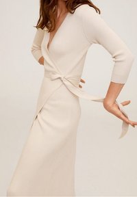 Femme portant une robe portefeuille en maille côtelée crème avec des manches trois-quarts, nouant la ceinture à la taille dans le dos.