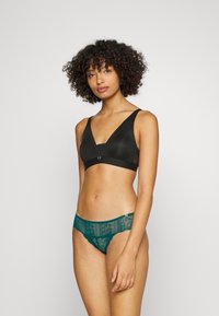 Czarny materiałowy biustonosz braletka z szerokimi ramiączkami i zapięciem z przodu, w komplecie z turkusowymi koronkowymi majtkami o skomplikowanych wzorach i falbaniastych krawędziach.