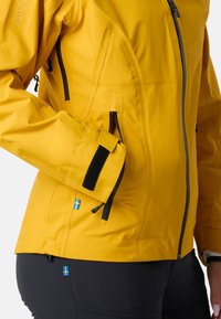 Gul vandtæt jakke med sorte lynlåse og accenter. Har flere lommer, justerbar manchet og blå svensk flag detalje.