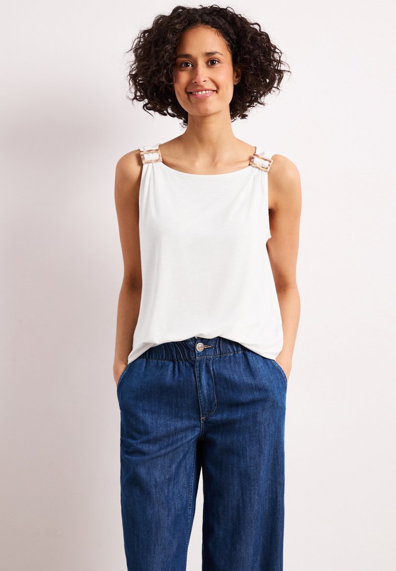 Femme souriante, portant un haut blanc sans manches avec des bretelles à boucle et un jean bleu ample taille haute, debout les mains dans les poches.