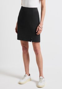 Jupe noire longueur genou en tissu doux, dotée d'une fente sur le côté et de deux poches. Portée avec des baskets blanches pour le contraste.