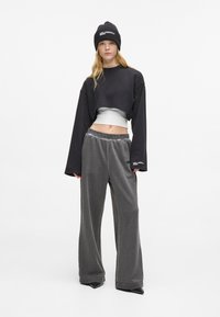 Ženska v črni zimski kapi, krajšem črnem puloverju preko belega vrha, širokih sivoh hlačah in črnih špicastih čevljih, stoji ob beli steni.