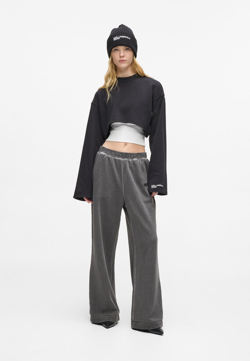 Ženska v črni zimski kapi, krajšem črnem puloverju preko belega vrha, širokih sivoh hlačah in črnih špicastih čevljih, stoji ob beli steni.