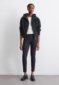Sudadera negra con cremallera y un acabado texturizado, combinada con leggings oscuros que tienen costuras laterales. Los mocasines negros completan el conjunto.