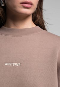 Felpa color marrone chiaro con colletto a coste, caratterizzata da testo bianco ricamato "WRSTBHVR" sul davanti in uno stile minimalista.