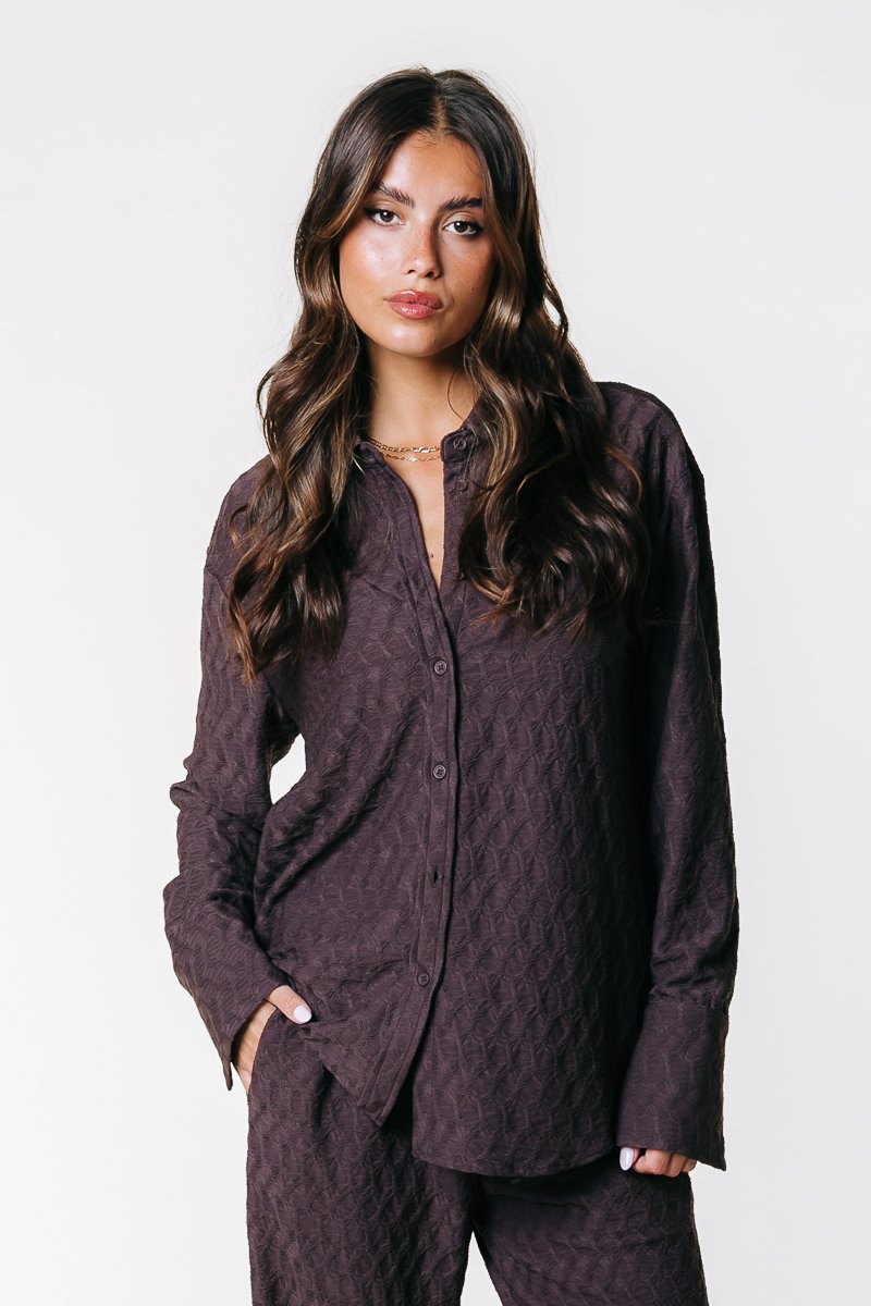 Camicia marrone a maniche lunghe con bottoni, realizzata in tessuto testurizzato con un motivo a rombi, indossata con pantaloni abbinati.