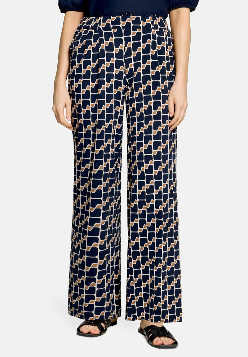 Femme portant un pantalon à jambes larges avec un motif géométrique bleu marine, beige et blanc, assorti à des sandales noires à bouts ouverts sur fond blanc.