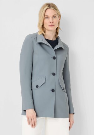 Frau trägt einen hellblauen, geknöpften Mantel mit Klappentaschen und steht vor einem schlichten grauen Hintergrund.