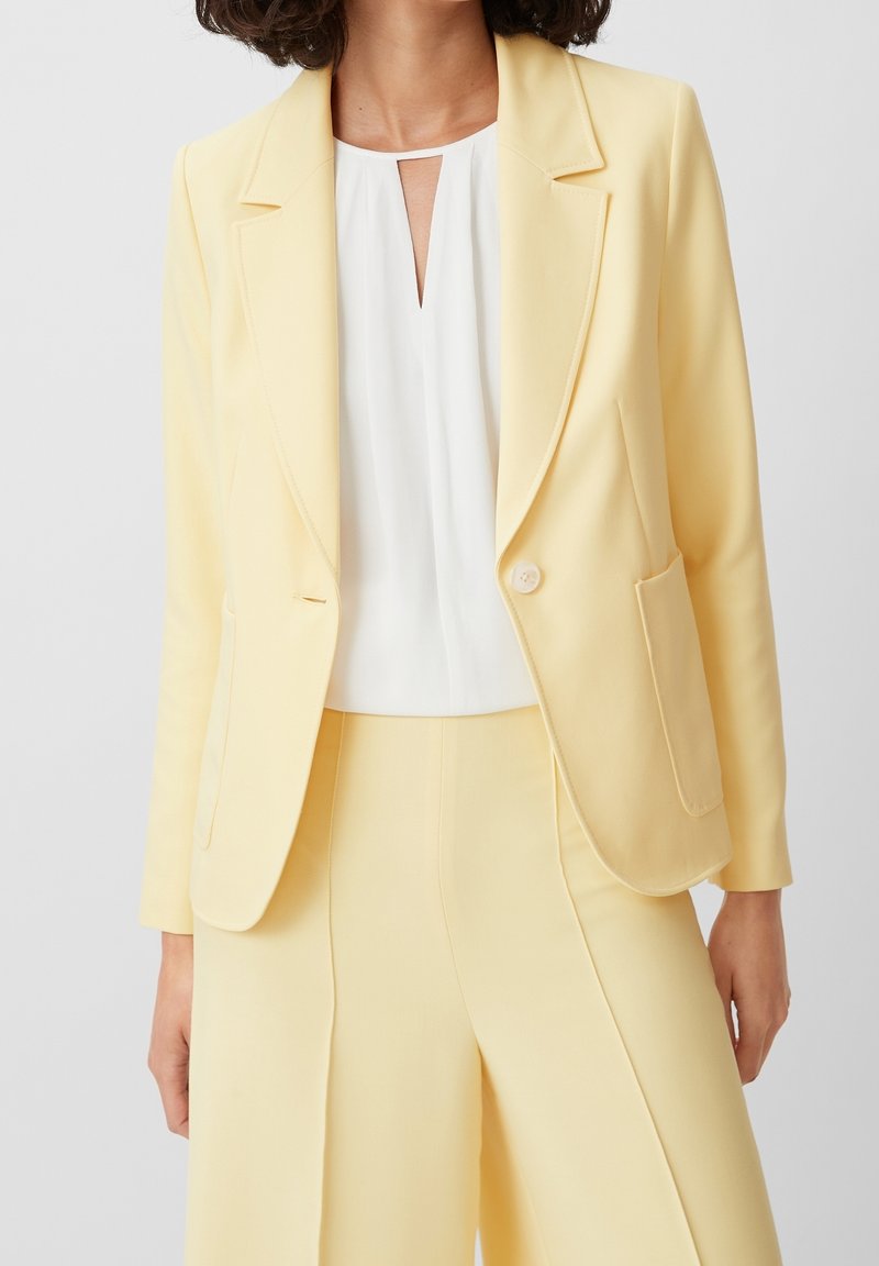 Femme portant un blazer jaune pâle et un pantalon assorti avec un chemisier blanc présentant une petite fente à l'avant.
