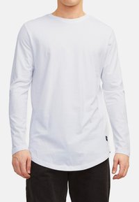 T-shirt blanc à manches longues en tissu doux avec un ourlet arrondi. Comprend un col rond et une petite étiquette logo sur le côté inférieur.